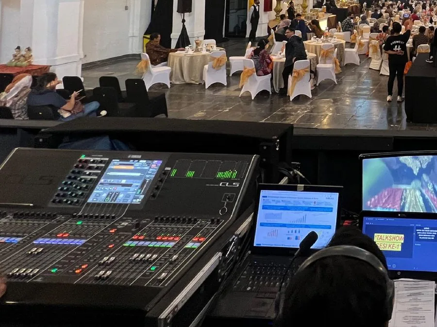 Meja kontrol dengan monitor multiview menghadap ruang seminar berisi tamu.